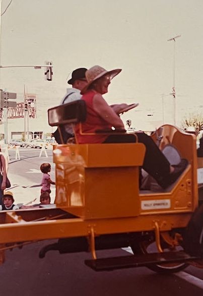 Two people in hats ride a vintage yellow vehicle in a parade, with bystanders watching from the roadside.