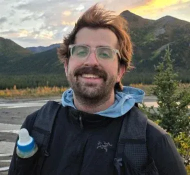 A person with a mustache and glasses smiles while wearing a jacket and backpack, outdoors with mountains in the background.