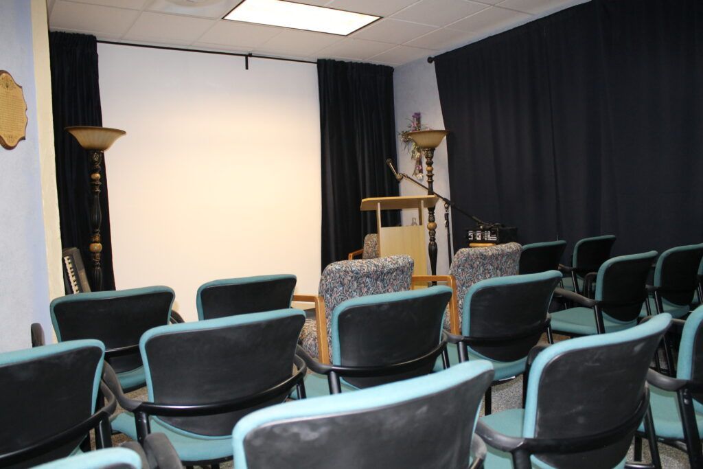 Small theater room with rows of blue chairs facing a white projection screen and stage curtains.