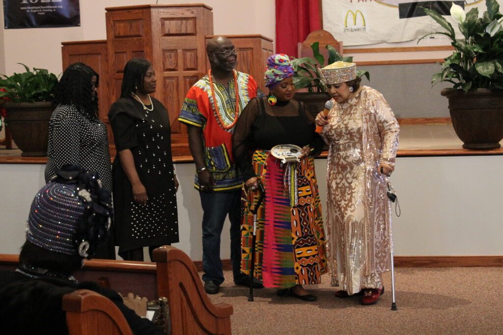 Five people standing on a church stage, one holding a cane, with wooden doors and potted plants behind them