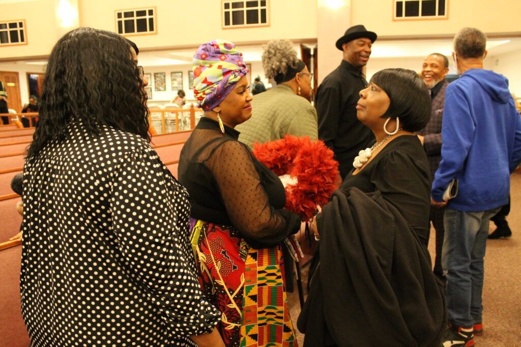 People chatting at an indoor gathering, with one holding bright red flowers and another in a colorful headwrap.