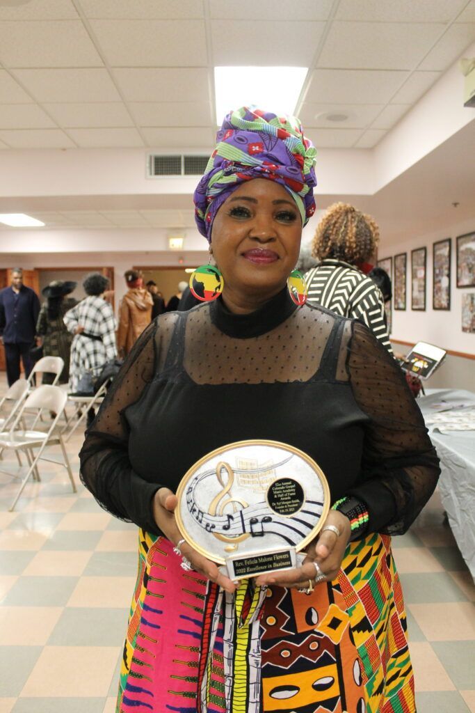 Woman in colorful headwrap and dress holding an award plaque in a hallway