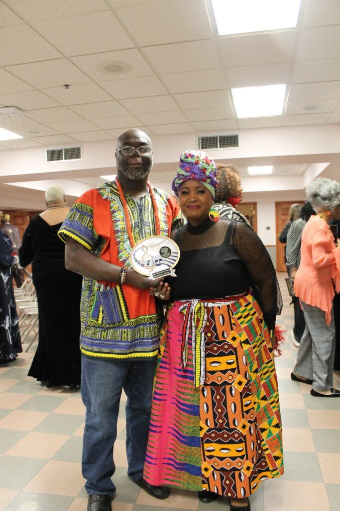 Two people in colorful patterned outfits standing together in a hall, holding a decorative plate