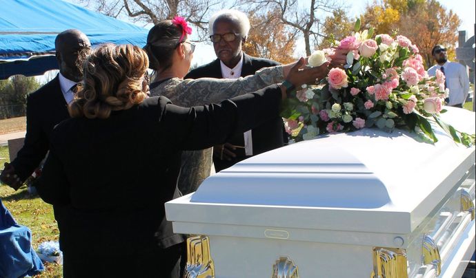 Mourners place pink flowers on a white casket at an outdoor funeral service.