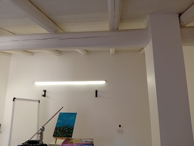 Stanza bianca e vuota con travi a vista, una lampada a parete e un piccolo cavalletto vicino al pavimento.
