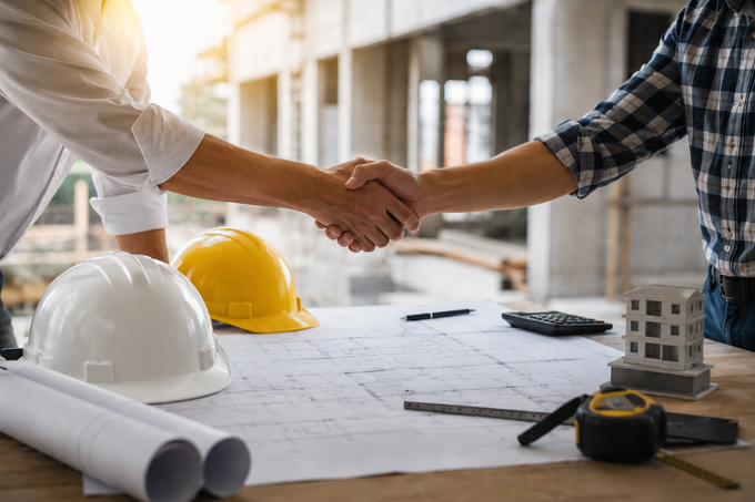 Due persone si stringono la mano in un cantiere edile, con progetti, caschi e attrezzi su un tavolo.