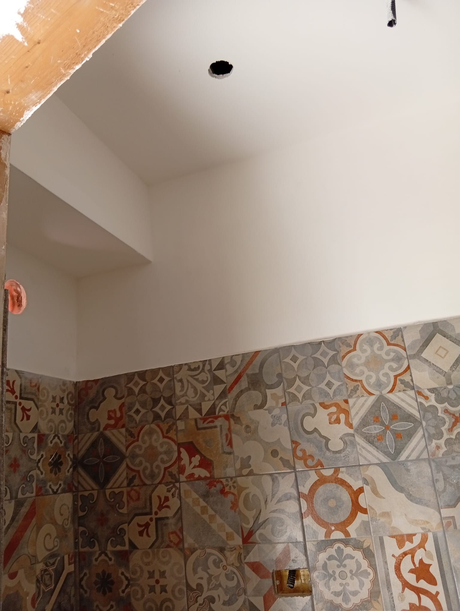 Parete del bagno parzialmente piastrellata con piastrelle decorate e apertura nel soffitto bianco non rifinito.