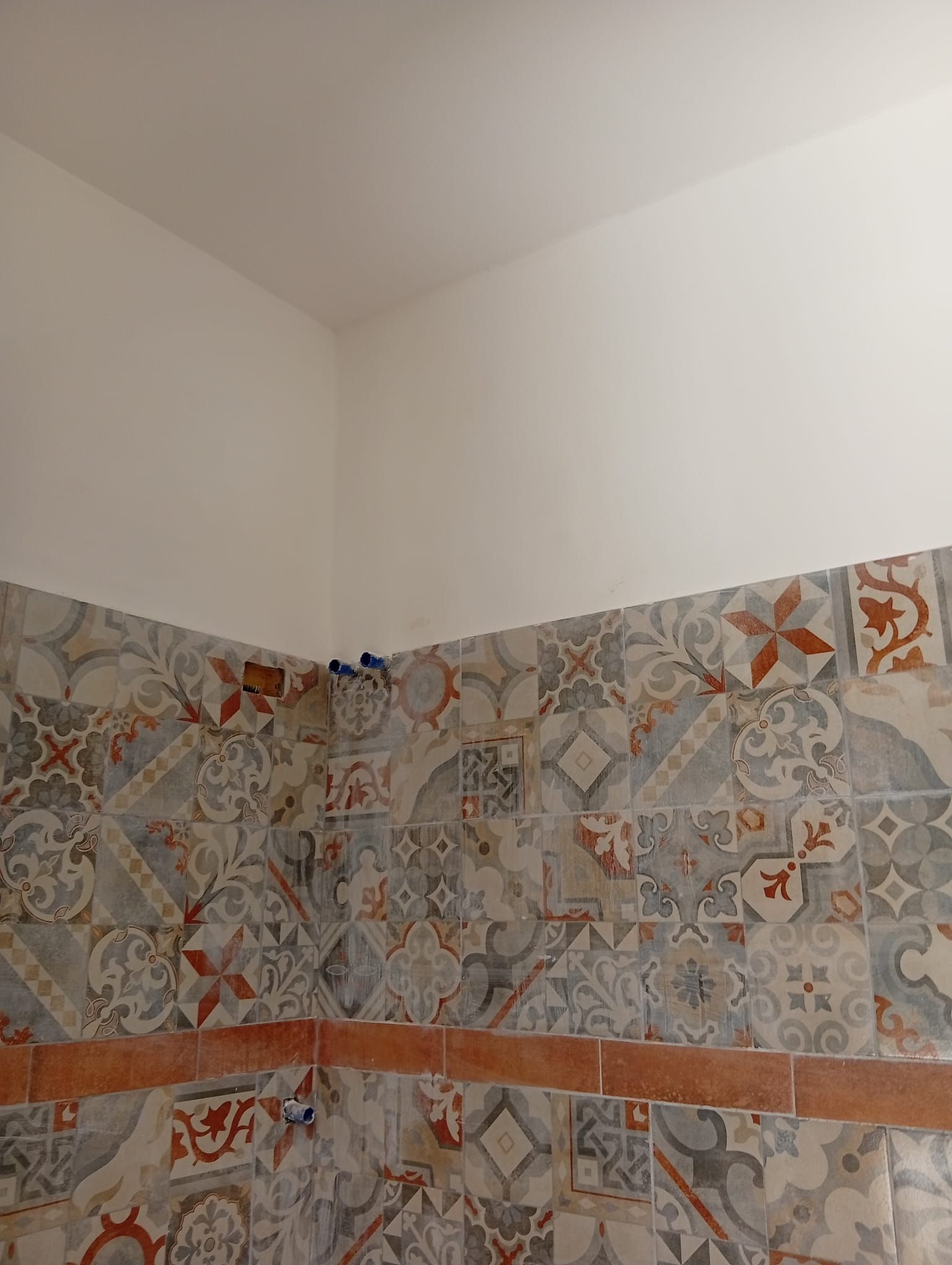 Angolo di una parete piastrellata del bagno con piastrelle decorate grigie, arancioni e beige sotto pareti bianche.