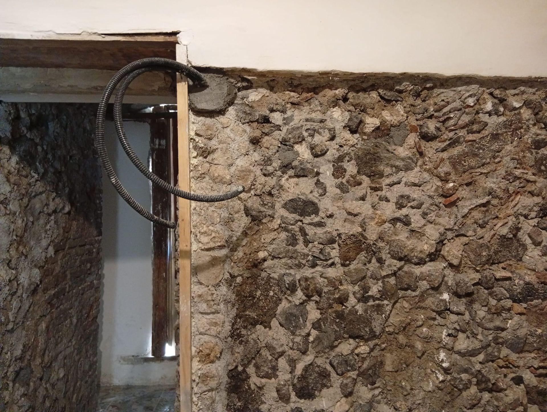 Muro danneggiato con barre d'armatura a vista e muratura in pietra grezza intorno a una porta.