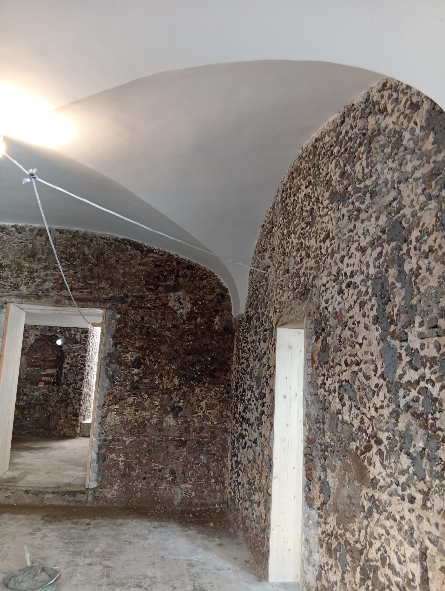 Stretto corridoio in pietra con soffitto a volta, pareti in pietra grezza, porte bianche e luce del sole proveniente da sinistra.