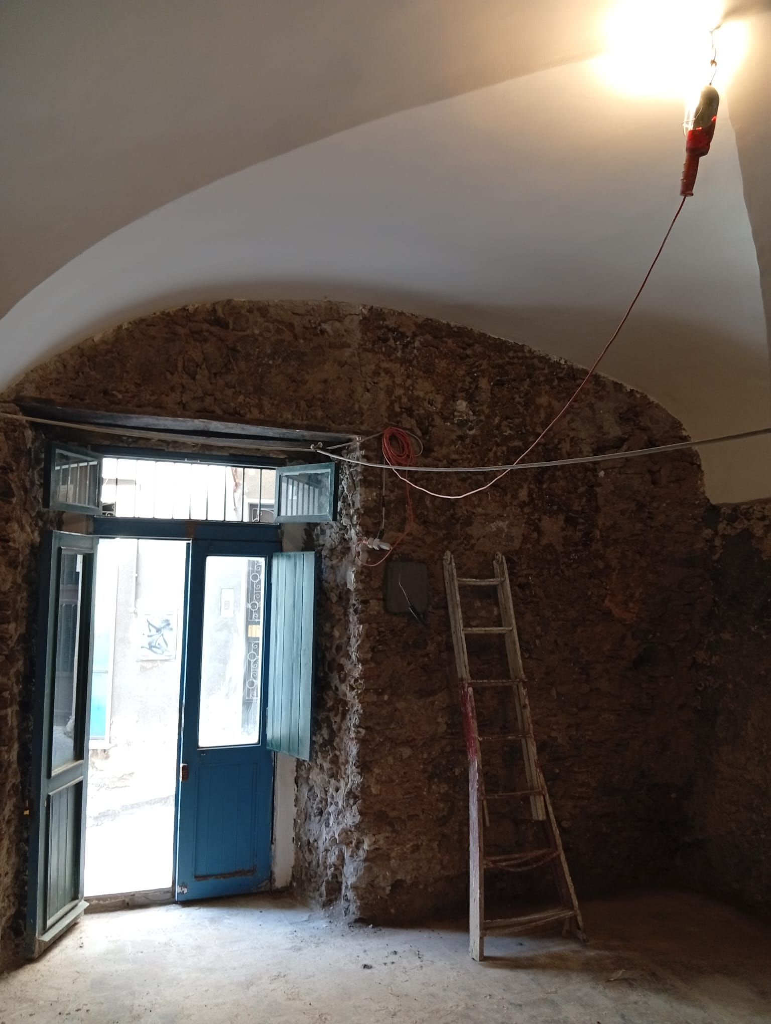 Vecchia stanza con pareti in pietra, soffitto a volta, porta blu aperta, scala e lampadina appesa.