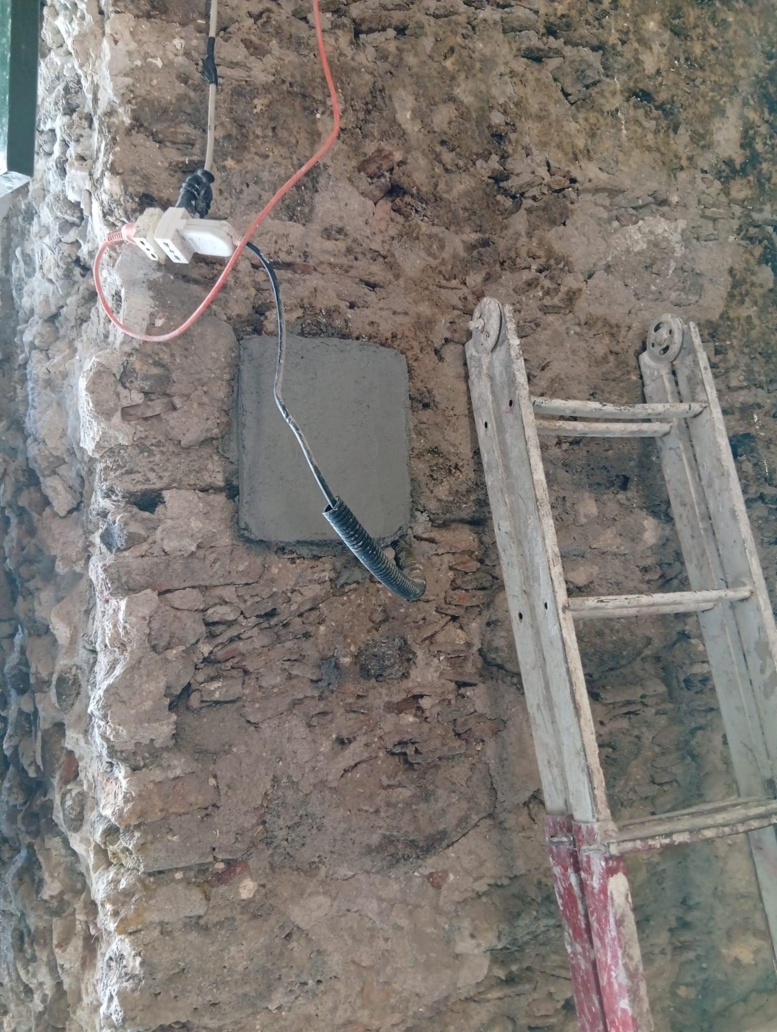 Muro di pietra grezza con una scala, lampadine appese e fili elettrici scoperti vicino a un piccolo pannello a muro.