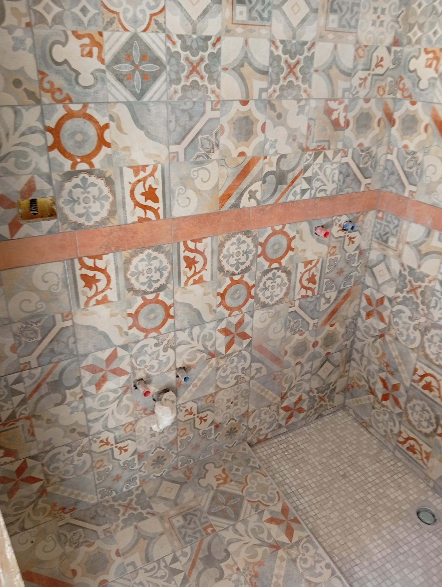 Doccia con piastrelle decorate con motivi geometrici arancioni, grigi e beige, rubinetto e scarico a pavimento.
