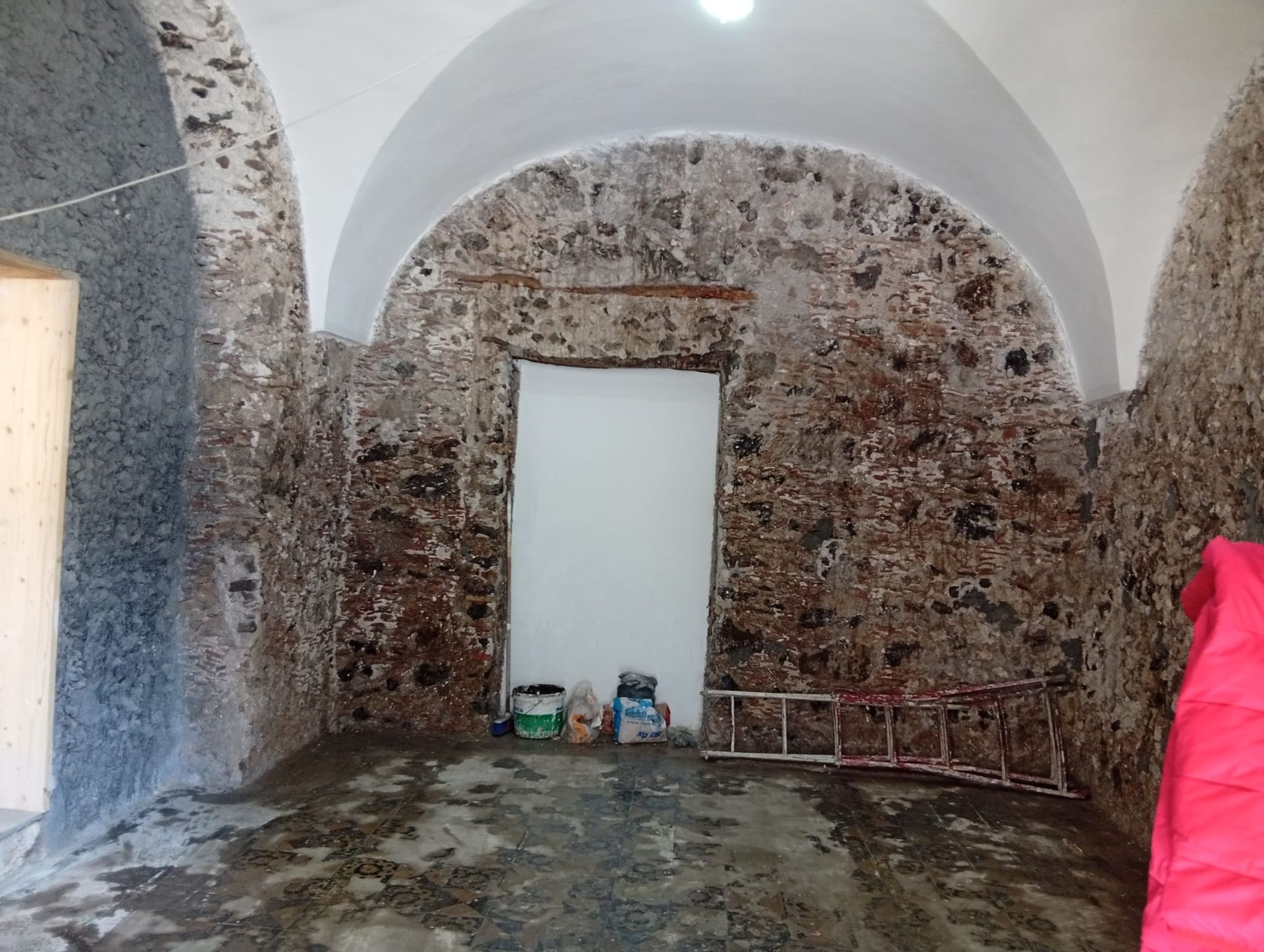 Stanza con pareti in pietra e soffitto a volta, porta bianca e attrezzi accatastati contro la parete di fondo.
