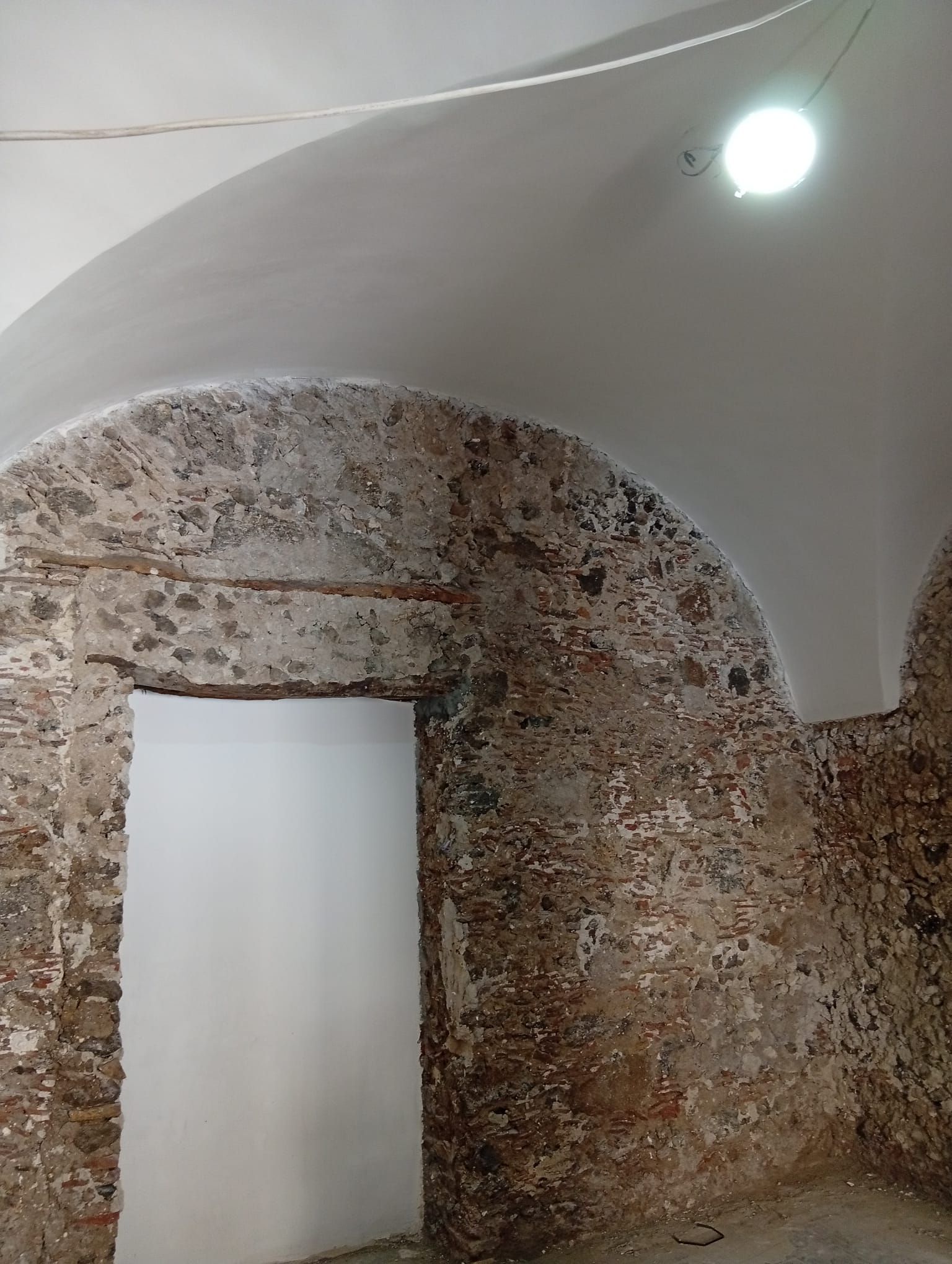 Stanza con pareti in pietra, porta ad arco e soffitto a volta bianco, illuminata da una lampada a soffitto.