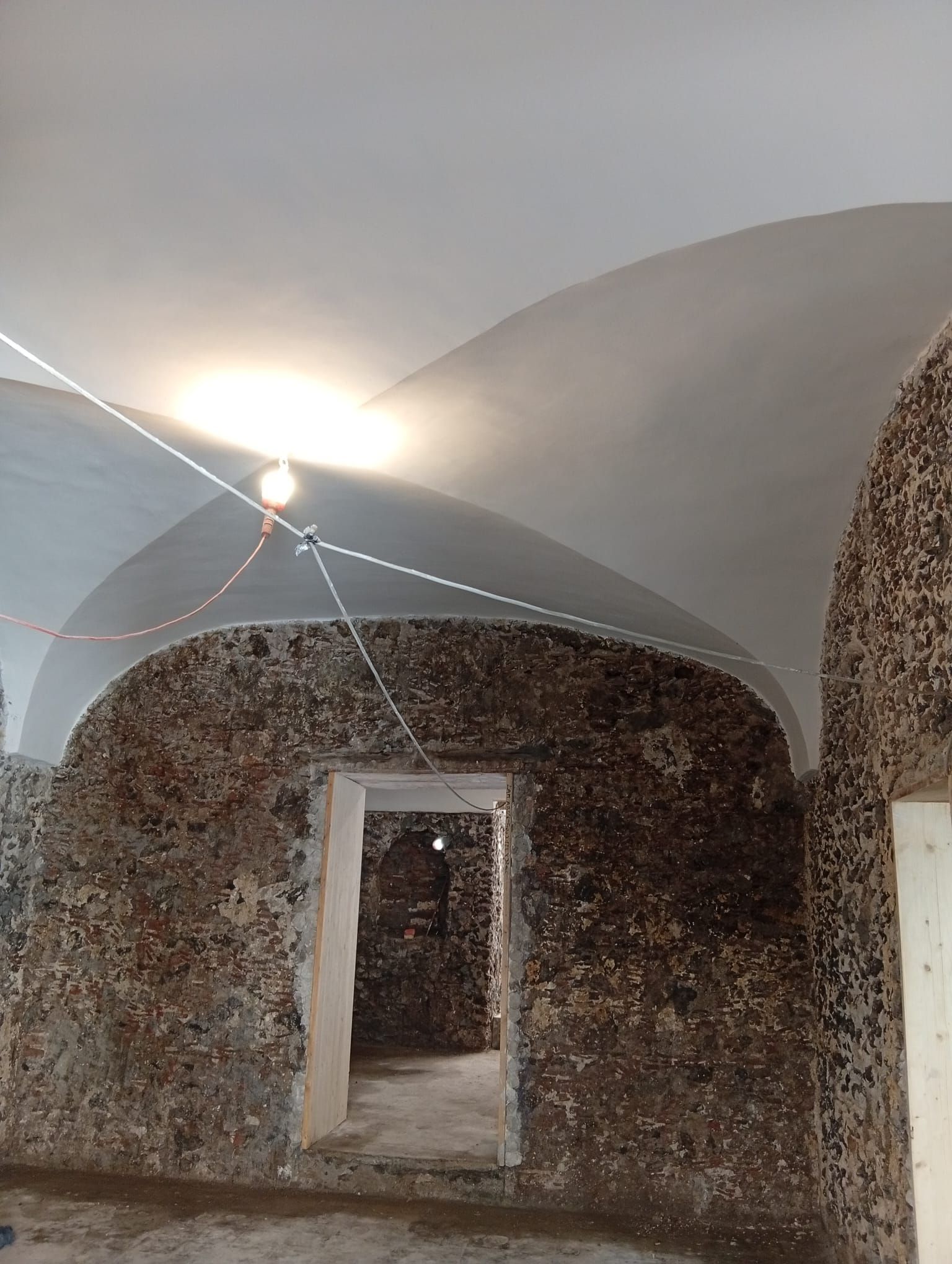 Stanza a volta con pareti in pietra, soffitto bianco e porta centrale che conduce a un interno in penombra