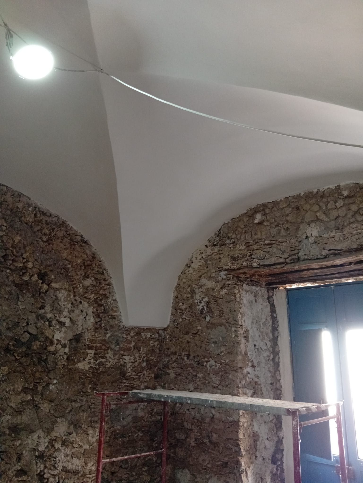 Interno di una stanza in pietra con soffitto a volta bianco, pareti a vista, una finestra e un'impalcatura sotto una lampadina nuda.