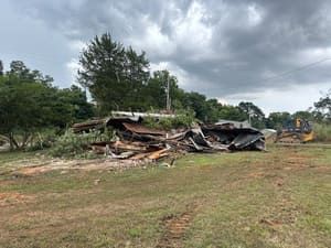 Collapsed building debris in a grassy field under cloudy skies, with a bulldozer in the distance.