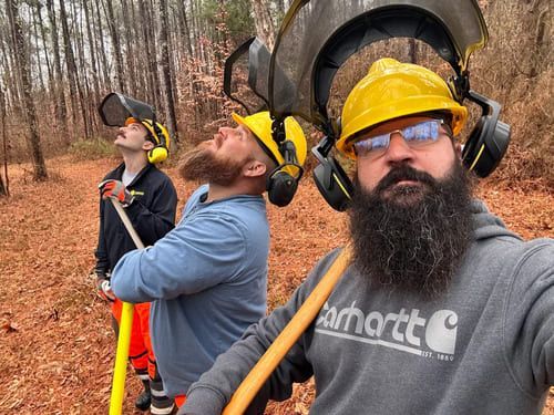 Three people in hard hats and ear protection at a muddy outdoor work site, one taking a selfie.