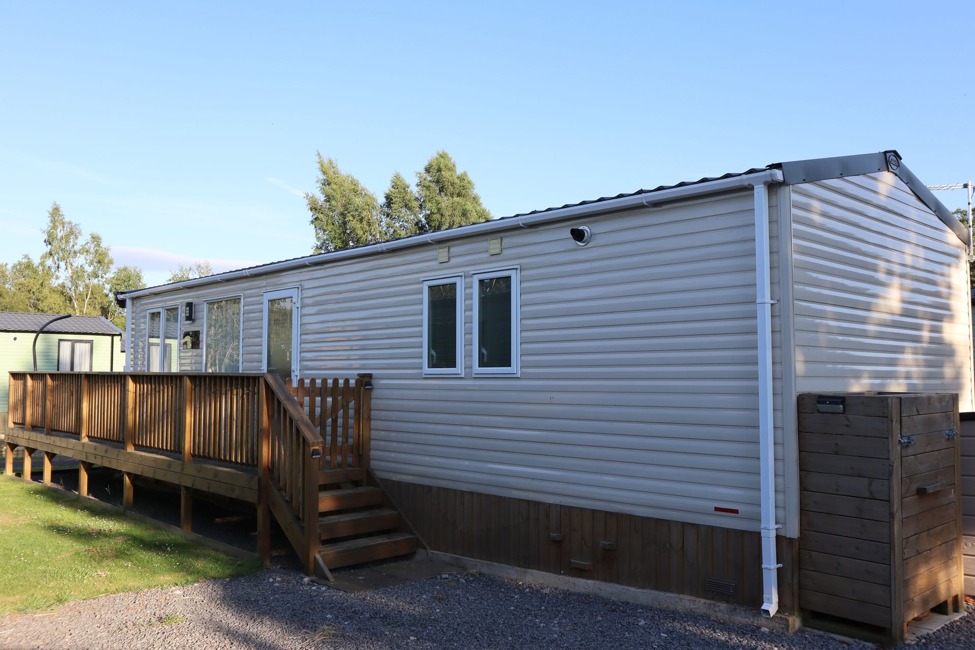 White mobile home with wooden deck and steps on a gravel lot under a clear sky
