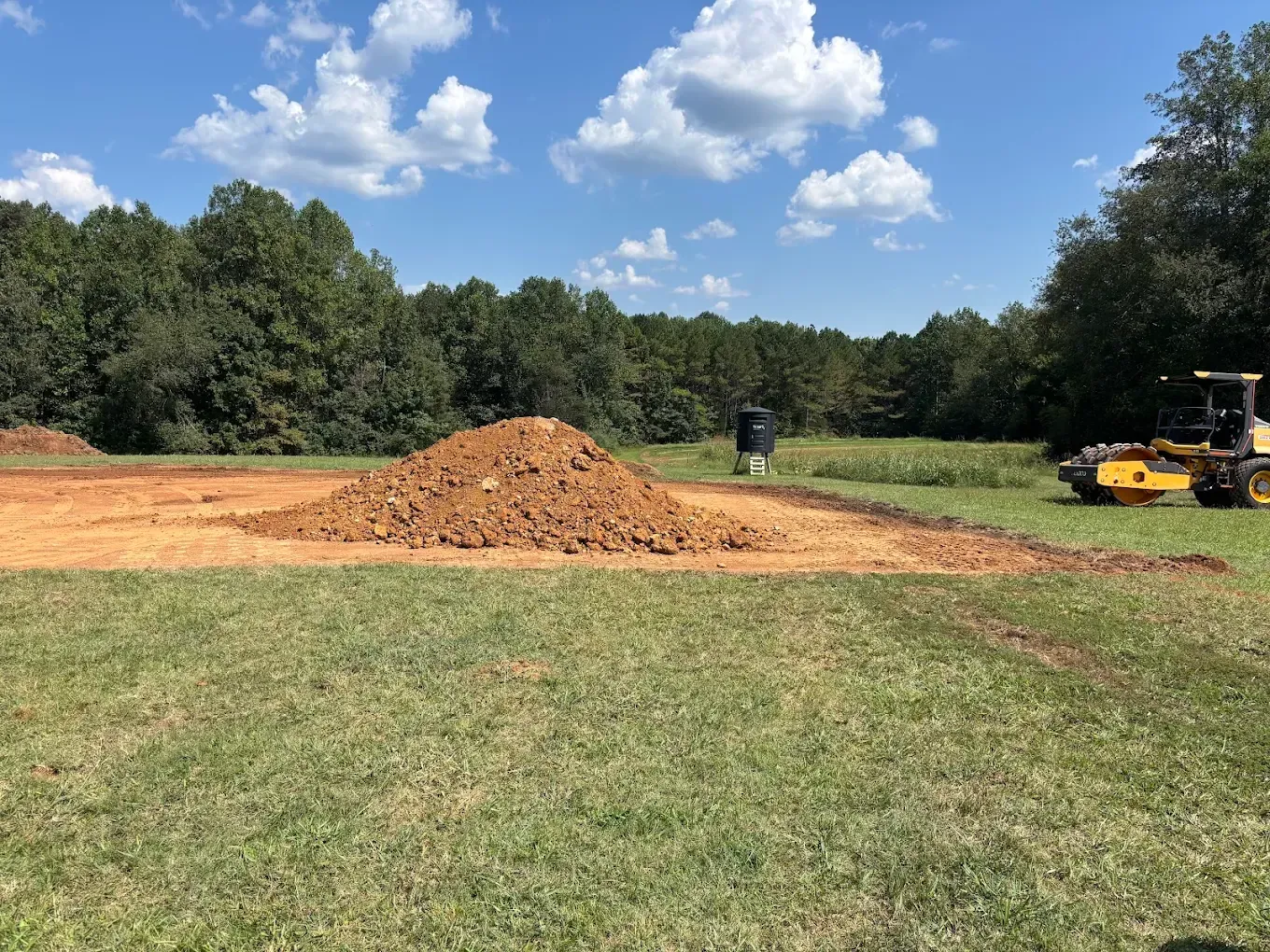 Grassy field with a dirt pile, worker, and yellow construction equipment under a blue sky