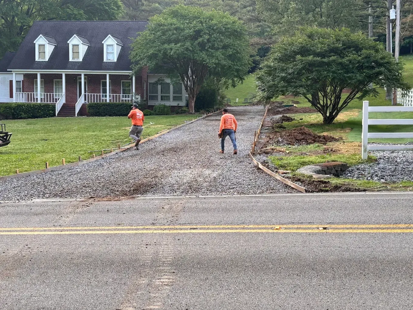Two workers in orange vests laying gravel on a driveway beside a house