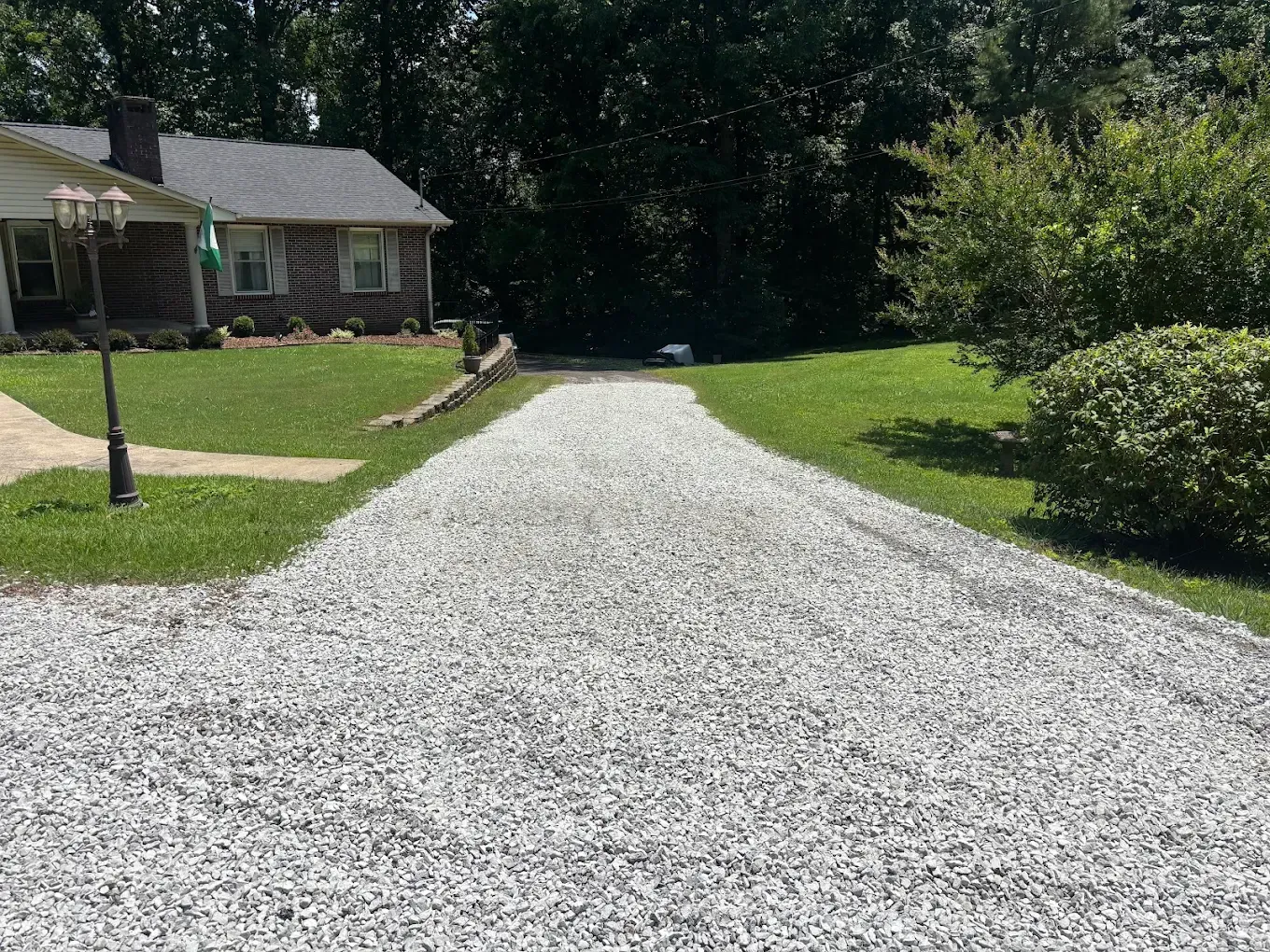 Gravel driveway leading to a house, bordered by green lawn and trees on a sunny day