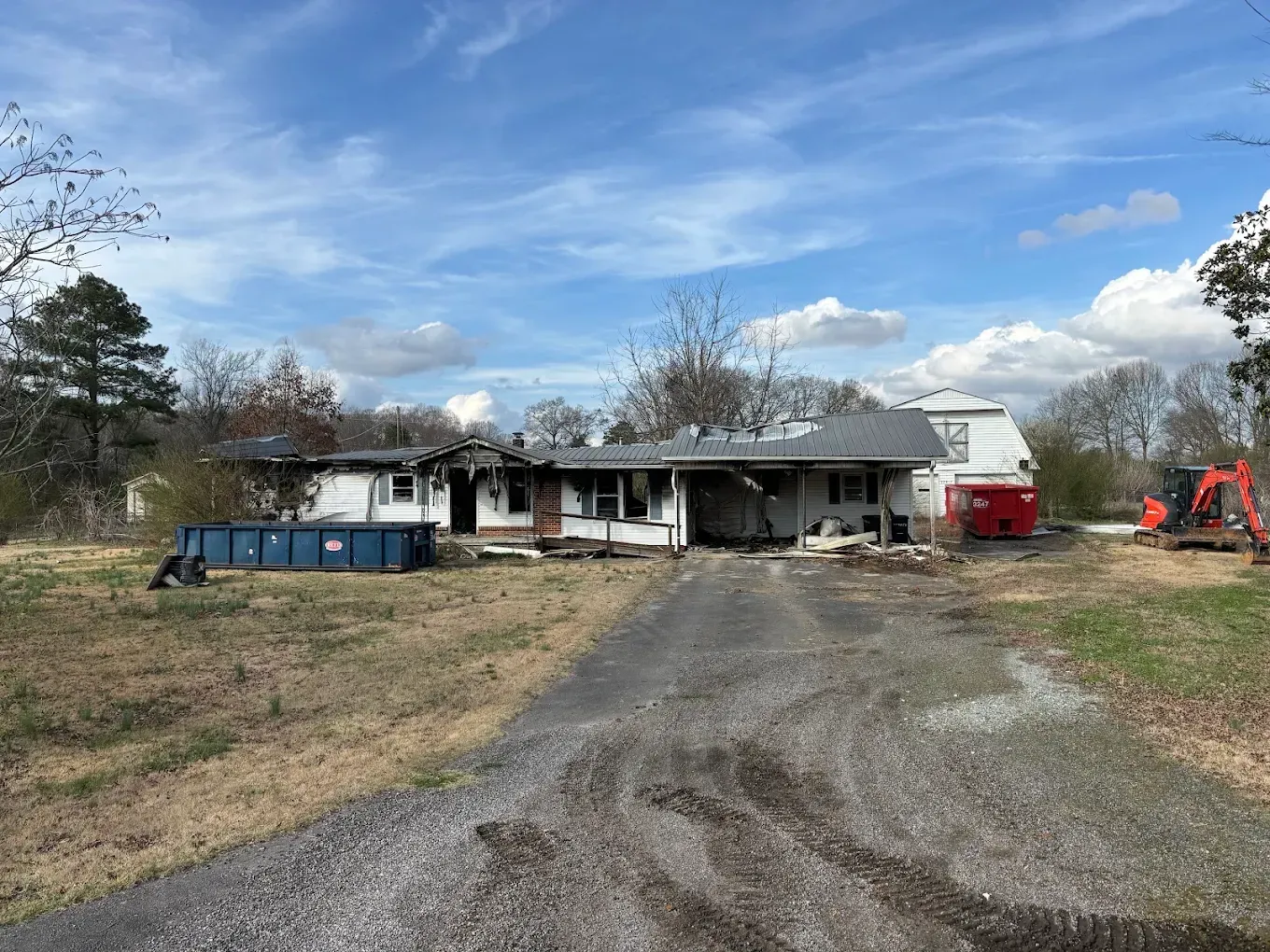 Rural house with gravel driveway, blue dumpster, and red excavator under a partly cloudy sky