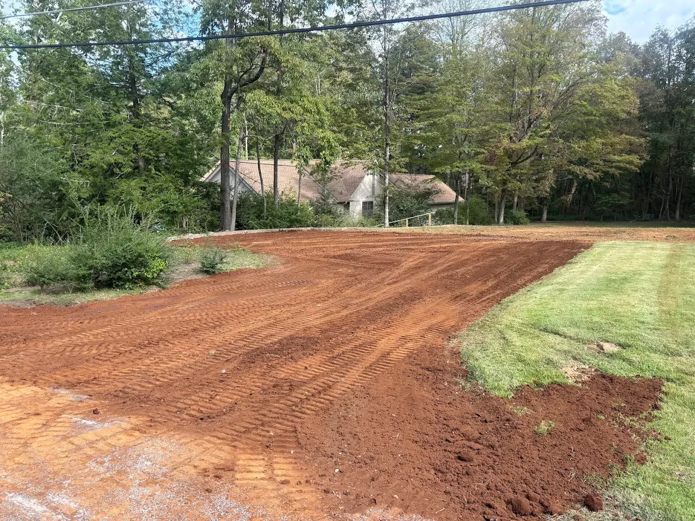 Freshly graded red dirt driveway beside a grassy yard and trees, with a house in the background