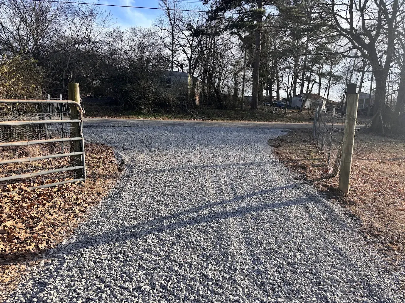 Gravel driveway entrance with a metal gate, wooden post, and trees along a rural road