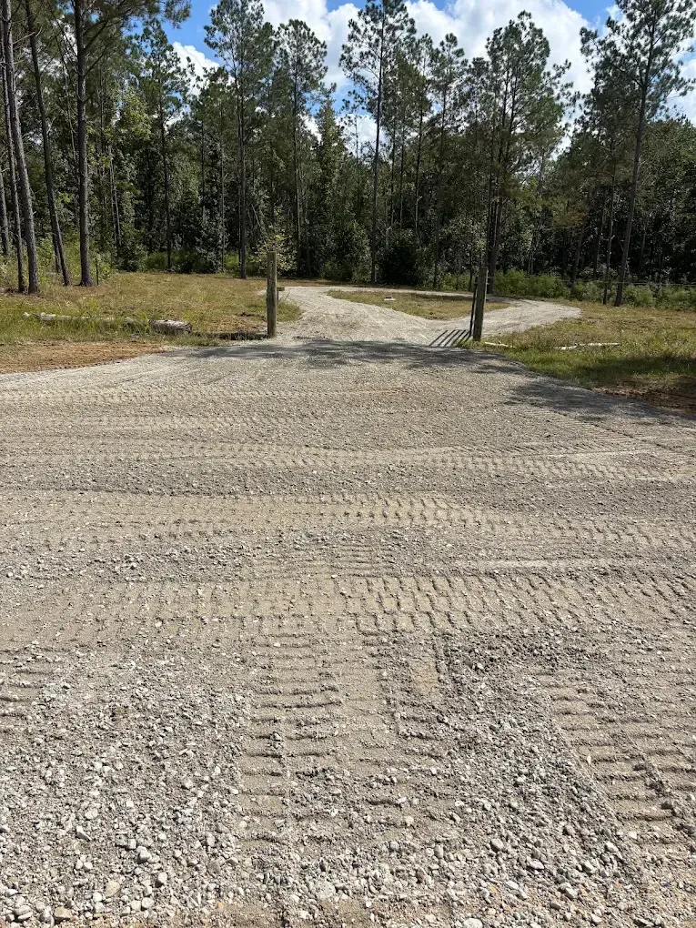 Gravel driveway with two wooden posts leading into a wooded area under bright daylight