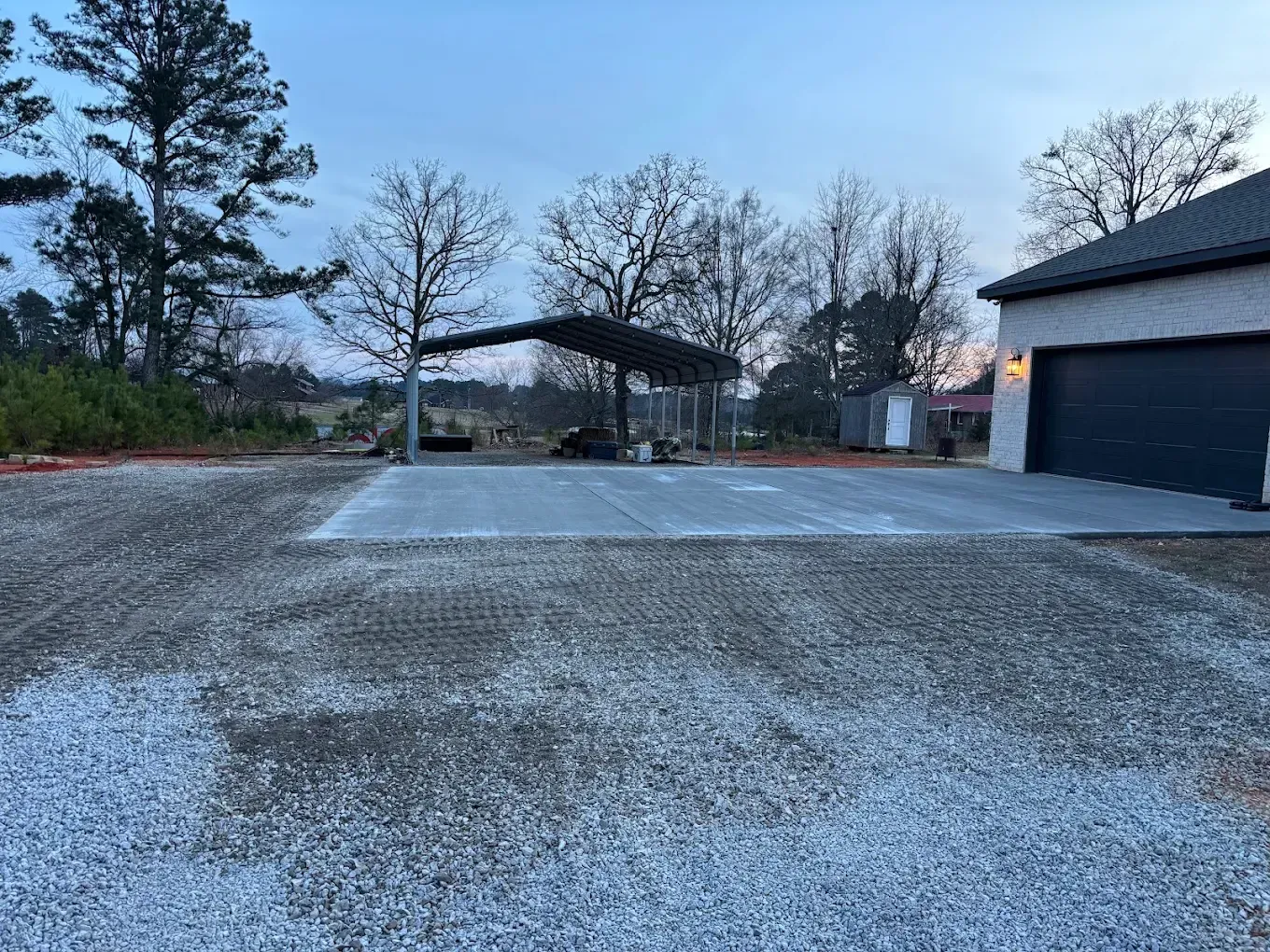 Gravel driveway beside a dark garage at dusk, with a carport and trees in the background.
