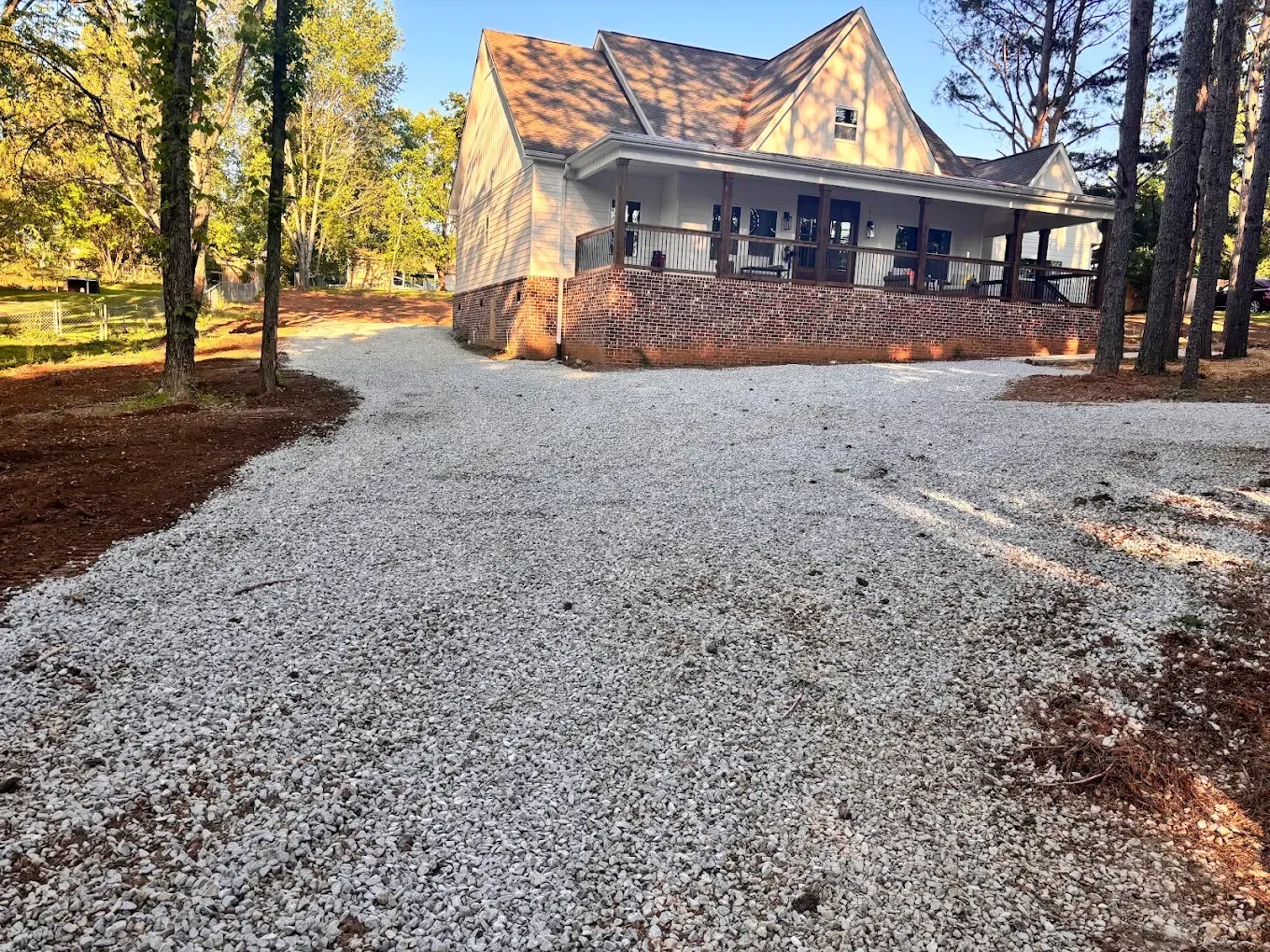 Gravel driveway leading to a white house with a porch, surrounded by tall trees in warm sunlight
