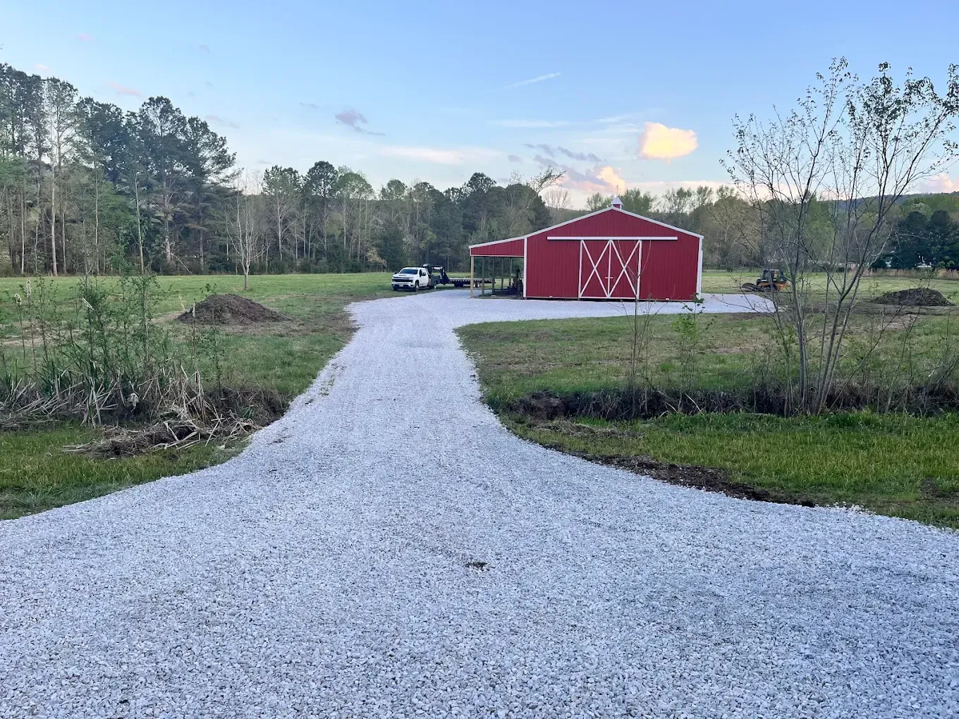 Gravel driveway leading to a red barn in a rural field with trees and a blue sky