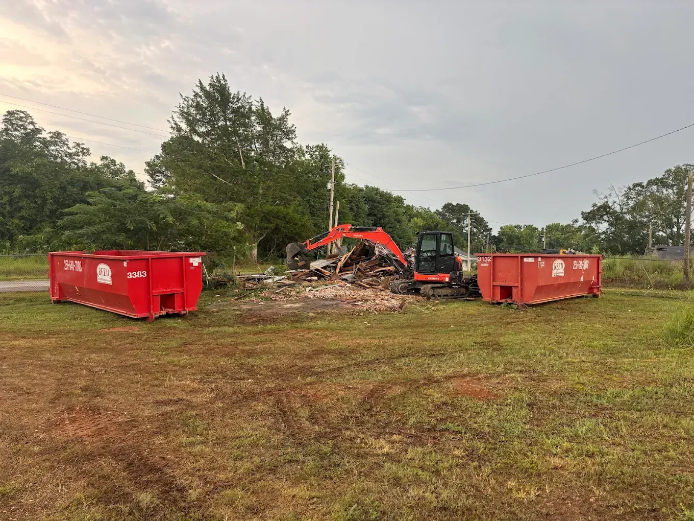 Excavator and red dumpsters beside a debris pile on a grassy lot with trees.