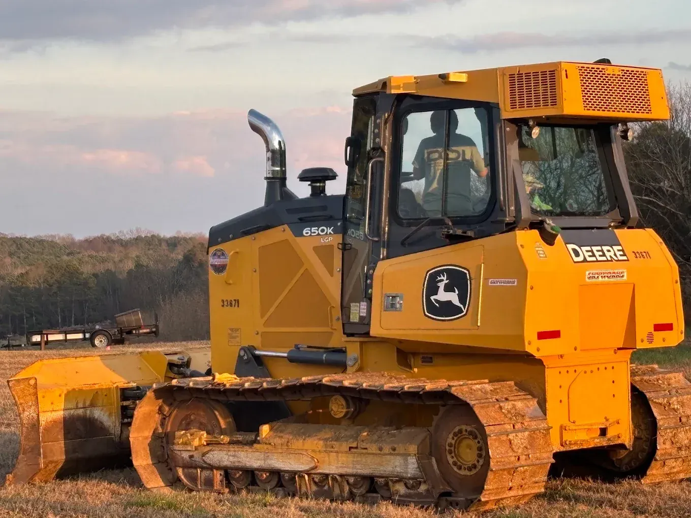 Yellow John Deere bulldozer on a dirt site at sunset, with blade lowered and tracks visible