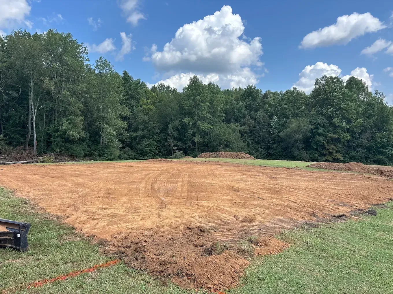 Cleared dirt lot bordered by trees under a blue sky with clouds