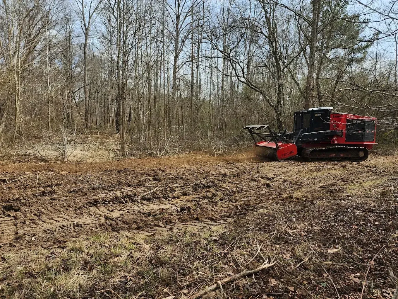 Red forestry mulcher clearing brush in a wooded clearing with bare trees.