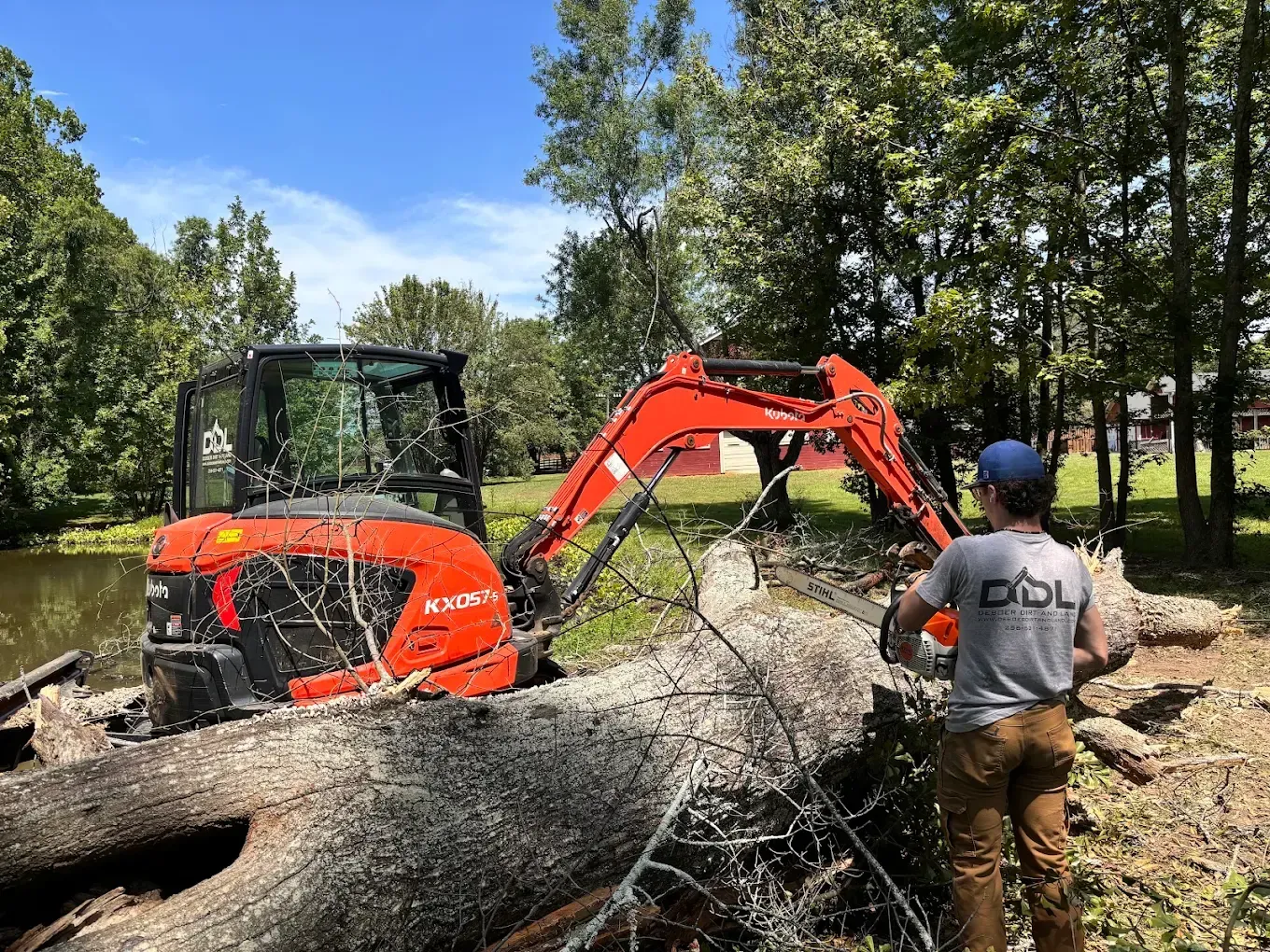 Orange excavator lifting a fallen tree beside a pond, with a worker standing nearby under trees