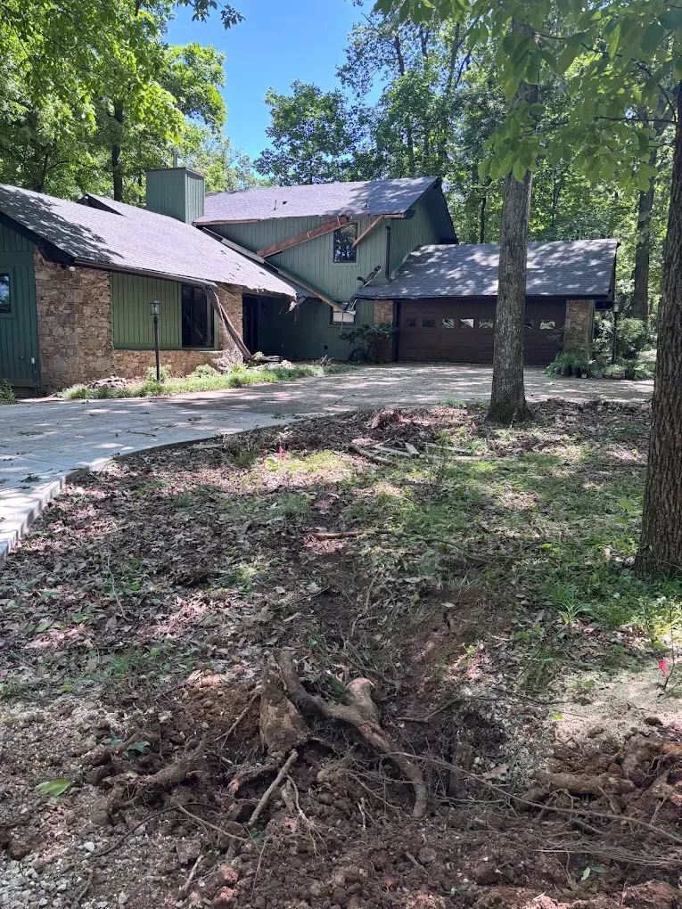 Stone house with dark roof beside a curved driveway, surrounded by trees and a grassy yard.