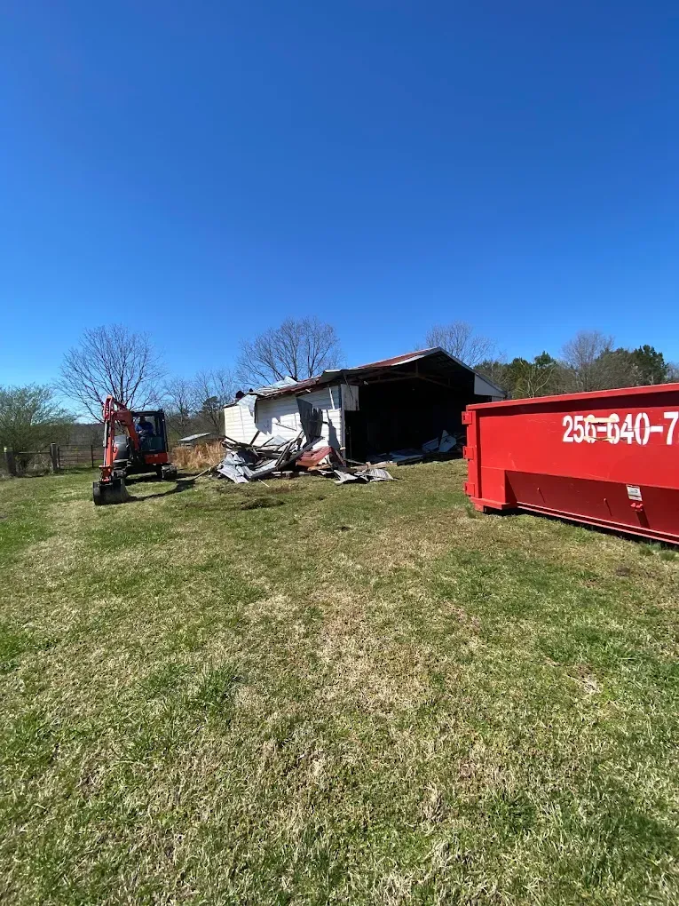 Damaged house with debris on a grassy hill, an excavator, and a red dumpster under a clear blue sky