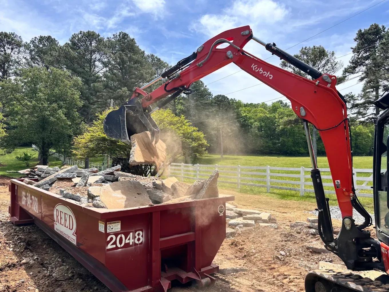 Red excavator dumping rock into a red dumpster in a wooded construction site under a blue sky.