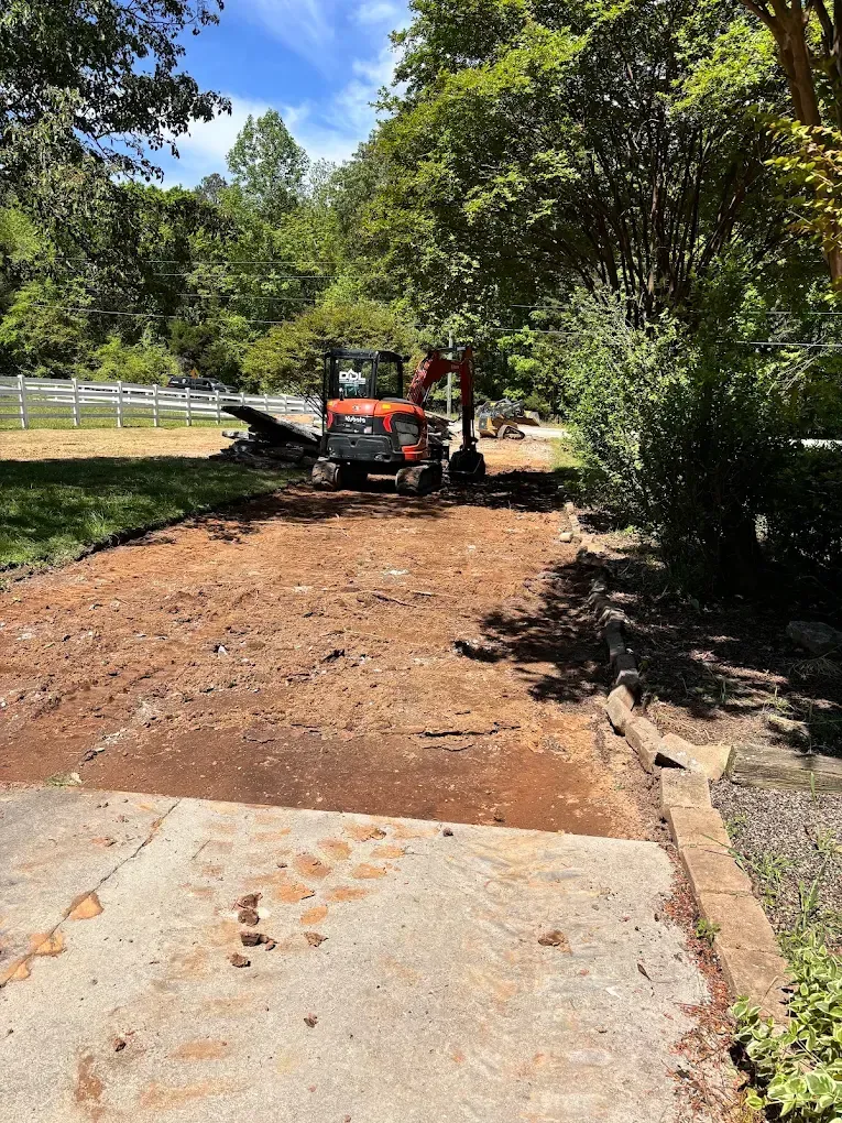 Orange mini excavator clearing dirt in a wooded yard beside a concrete slab.