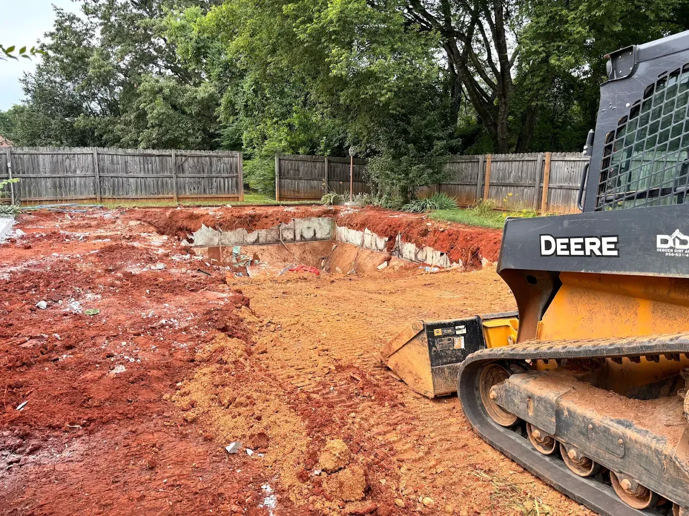 Orange excavator digging a backyard excavation site with red dirt and a wooden fence