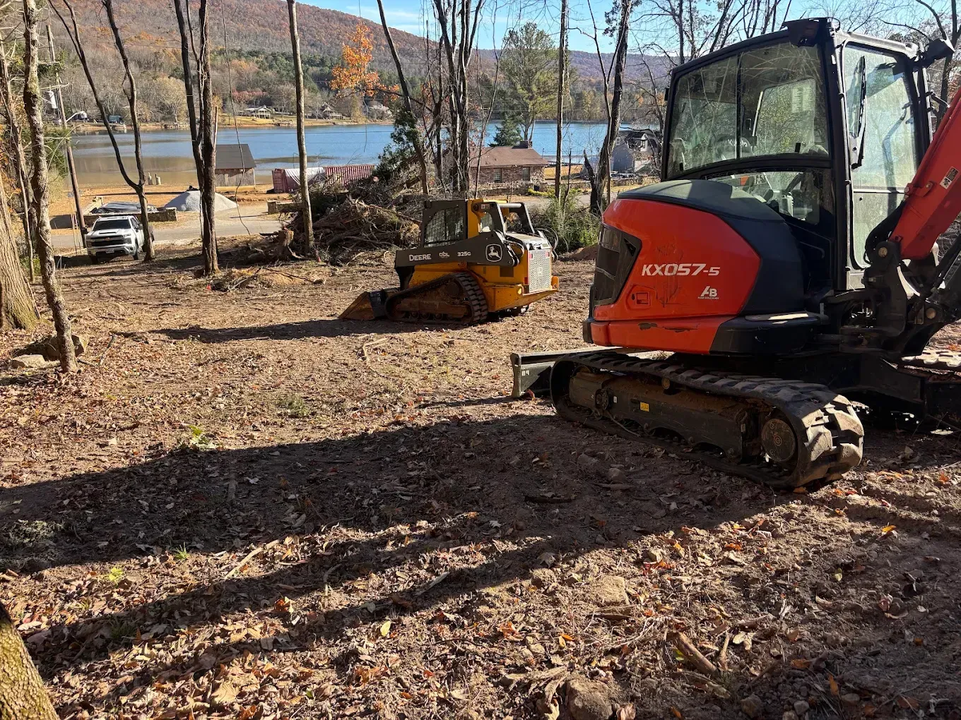 Red excavator on a leaf-covered lot near a lake with autumn trees and mountains in the background