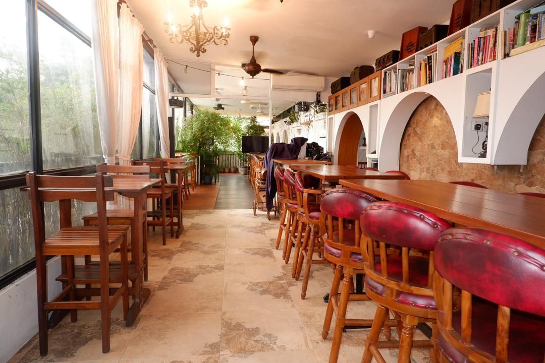Cafe interior with wooden tables, red leather chairs, and a bookshelf in an archway.