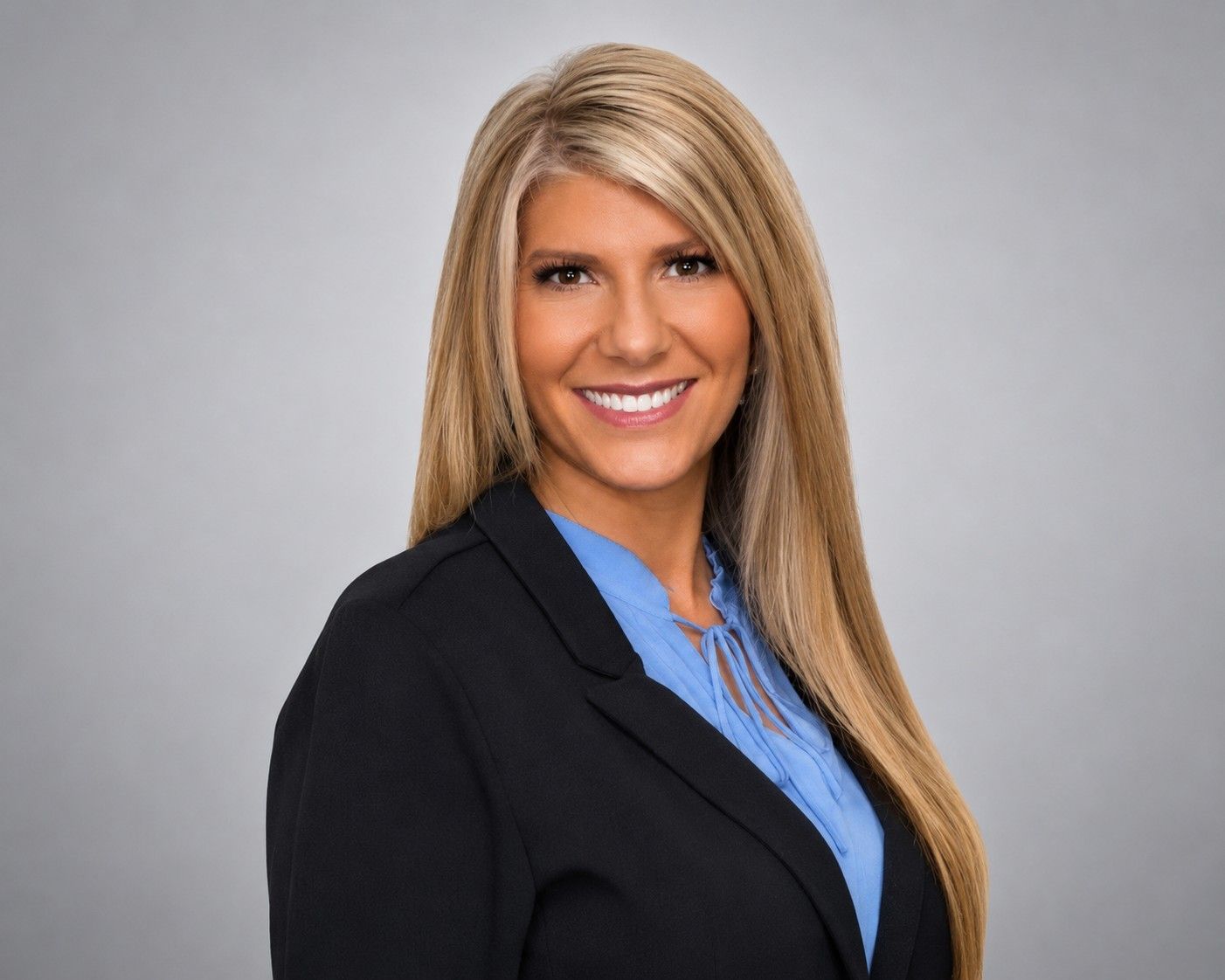 Professional headshot of a smiling person in a black blazer and blue shirt against a gray background
