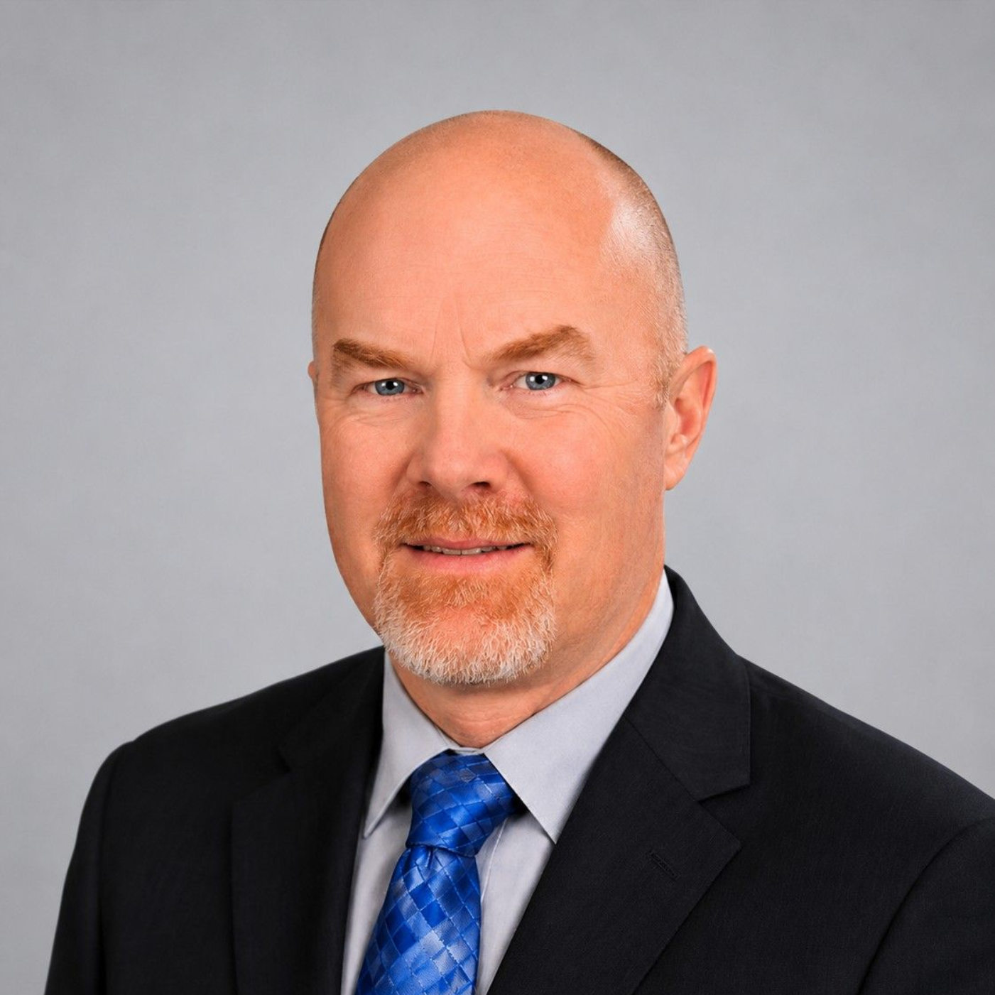 Professional headshot of a bald man in a black suit, gray shirt, and blue tie against a blurred blue background