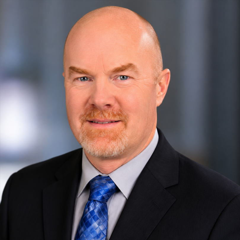 Professional headshot of a bald man in a black suit, gray shirt, and blue tie against a blurred blue background