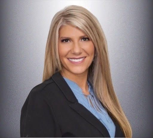 Professional headshot of a smiling person in a black blazer and blue shirt against a gray background